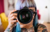 Woman with purple hair aiming a camera directly at the viewer